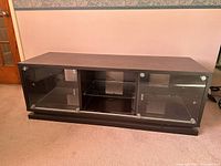 Front view of the black TV stand showing wood structure and transparent sliding glass doors with metal knobs, placed on a beige carpet.