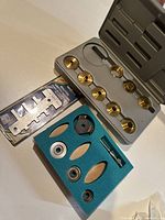 Gray plastic case containing nine brass bushings arranged in two rows. Accompanying blue foam insert holds various metal parts including bearings and a collet-like piece. Metal router depth gauge visible in original Samona packaging alongside the case.