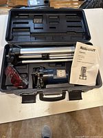 Carrying case open showing laser level, tripod folded, glasses, and manual inside.
