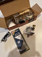 Opened box containing various metal tool accessories including clamps and adjustment parts packaged loosely.