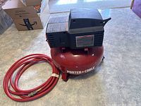 Lateral view of red Porter-Cable air compressor with attached red air hose coiled beside it on a floor surface.
