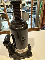 View of black hydraulic jack with threaded screw lift and base plate on white surface, showing general condition and structure.