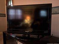 Front view of Samsung 32 inch LCD TV showing black frame, pedestal stand, and reflected light from room.