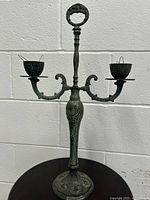 Full view of antique brass or bronze two-arm candelabra with patina, standing on a round decorative base with floral patterns, against a white brick wall.