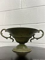 Full side view of antique bronze handled bowl with green patina on a dark surface against white brick wall.