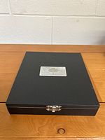 Closed black storage box for the cheese cutter set with silver emblem on top.