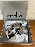 Photo of open silver box showing pewter synthetic high heel sandals inside wrapped in tissue paper