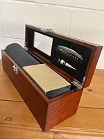 Wooden box open, showing foam lining and two silver wine accessories in black foam insert along with beige booklet.