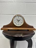 Wood framed mantel clock on a black wooden stool against a white brick wall. Shows full clock body and shape with visible wood grain and finish.