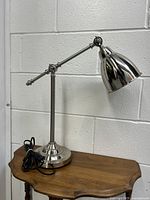 Full view of the chrome metal desk lamp showing its adjustable arm, pivoting lamp head, circular base, and corded power cable.