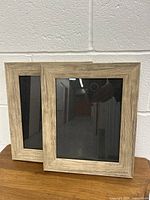 Two wood-toned plastic photo frames side by side on a wooden surface against a white brick wall.