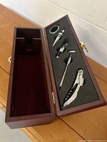 Open view of the red brown wooden wine gift box showing the foam insert with five wine accessories inside: corkscrew, thermometer, two bottle stoppers, foil cutter.