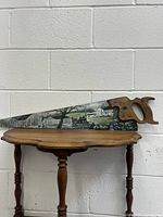 Full view of vintage decorative hand saw resting on a wooden table against a white brick wall background showing entire blade and handle with painted artwork.