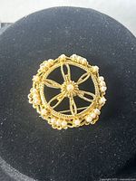 Top view of the gold tone filigree brooch with faux seed pearls on a black display stand.