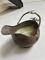Side angle of vintage brass coal scuttle showing the curved spout, porcelain handles with blue Delft floral pattern, and lion head hinges.