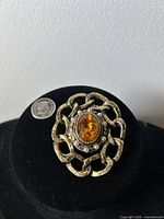 Front view of vintage brooch with gold-tone filigree and central amber-colored stone beside a Canadian quarter for size reference.