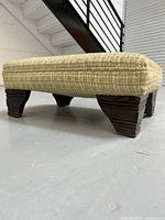 Side angled view of footstool showing beige woven textured upholstery and dark wooden legs with stacked design.
