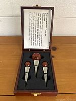 Top-down view of open wooden box containing three wine bottle stoppers with rounded wooden tops and metal tapered bodies. Included user instructions printed on the inside of the lid.