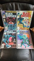 Four Black Panther comic books arranged in two rows on a black chair, covers visible with various cover art.