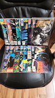 Photo showing front covers of 14 vintage Detective Comics issues arranged in two rows on a black chair. Covers show colorful comic book art, many featuring Batman.