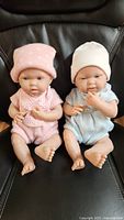 Two JC Toys La Newborn realistic baby dolls seated side by side on black chair, one in pink outfit and hat, the other in blue outfit and cream hat.