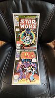Two Marvel Star Wars comic books in protective sleeves on black chair, showing full covers with iconic Star Wars characters.