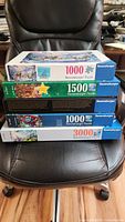 Five boxed Ravensburger puzzles stacked on a chair showing box front and side: two 1000-piece puzzles, two 1500-piece puzzles including Christmas edition, one 3000-piece puzzle.