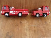Photo showing side profile of two red Buddy L fire engine toy vehicles placed on wood background; larger vehicle on left, smaller on right; vehicles have intact plastic windows and detailed fire truck decals