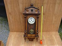 Front view of the antique wooden regulator clock mounted against a wooden panel, showing overall design and size with measuring tape for scale.