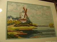 Cross stitch artwork showing a detailed windmill and landscape scene under glass in a wooden frame.