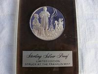 Photo of the sterling silver proof coin in protective case showing a scene of children and adults around a campfire, with label reading 'Sterling Silver Proof Limited Edition Struck at The Franklin Mint'