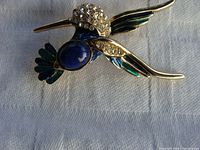 Top front view of the hummingbird brooch showing rhinestones on the head and gold-tone metal with blue cabochon and enamel accents.
