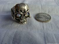 Front view of silver skull ring next to coin for size reference. Displays iridescent rhinestone eyes and detailed skull features.