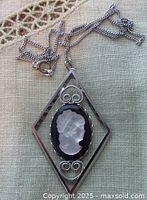 Top view of the diamond shaped silver tone cameo pendant on chain lying on lace cloth showing the intricate cameo detailing and chain clasp.