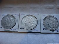 Four US Morgan Silver Dollar coins in protective holders showing obverse sides with the dates 1889, 1885, 1895, and 1891 visible, some wear and blemishes including what looks like a BB shot mark on one coin.