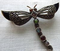 Front view of vintage sterling silver dragonfly brooch with detailed openwork marcasite wings, black enamel tips, and multi-colored round gemstones along the body including red, green, blue, and pink stones. Two red stone eyes and square amethyst in center.