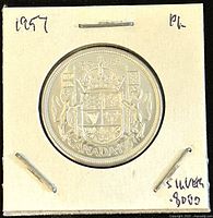Reverse side of the 1957 Canadian 50 cents coin showing the Canadian coat of arms with inscription 'CANADA 1957'. Coin is in a cardboard holder stapled around the edges and marked 'PL' and 'Silver 80%'.