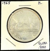 Reverse view of 1965 Canadian silver dollar coin showing canoe with voyageurs and date 1965, with silver content stamped on holder.