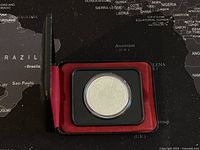 Photo of the closed black hinged display case with red lining, showing the coin inside a transparent rectangular plastic holder.