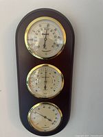 Wooden plaque holding three circular dials: top thermometer, middle hygrometer, bottom barometer with gold bezels.