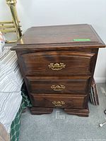 Front view of a wooden bedside table showing three drawers and brass handles, placed next to a bed.