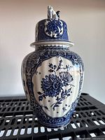 Full front and side view of the tall blue and white Delfts Boch ceramic lidded jar showing floral pattern and sculpted lid finial.
