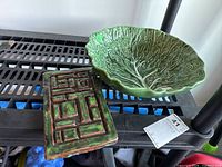 Rectangular green and brown glazed pottery plaque with geometric textured pattern, shown beside cabbage leaf bowl on a shelf.