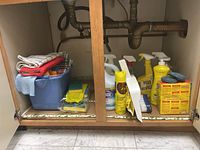 Full view of cleaning supplies under the sink, showing various bottles, SOS pad box, blue bucket, folded tea towels, and dustpan with broom.