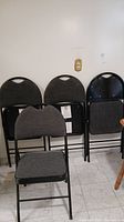 Four folding chairs, three folded and one unfolded, showing charcoal grey fabric and black metal frame.