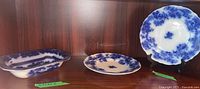 Three items: one oval-shaped W. Adams Shanchai bowl on left, two round Flow Blue china plates center and right, with one plate on stand showing shape.