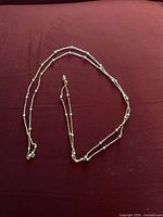 Gold tone ball chain necklace with small round beads along the length, laid out on a dark red fabric background.