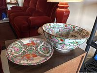 Hand painted Asian bowl and matching plate displayed on wooden surface with red couch and lamp in background, showing size and shape