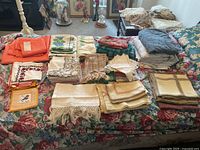 Overview of all linens showing various folded tablecloths, napkins, and bedsheets in different colors and patterns arranged on a floral covered couch and table.