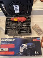 Black plastic case containing red Samona rotary tool with power cord and multiple accessory heads, box for Mastercraft Multi-crafter Tool Kit underneath showing wear